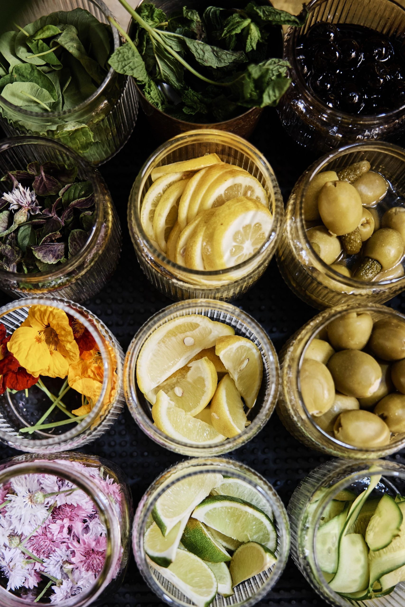 An overhead view of several jars filled with garnishes, including herbs, lemon slices, olives, edible flowers, and lime wedges.