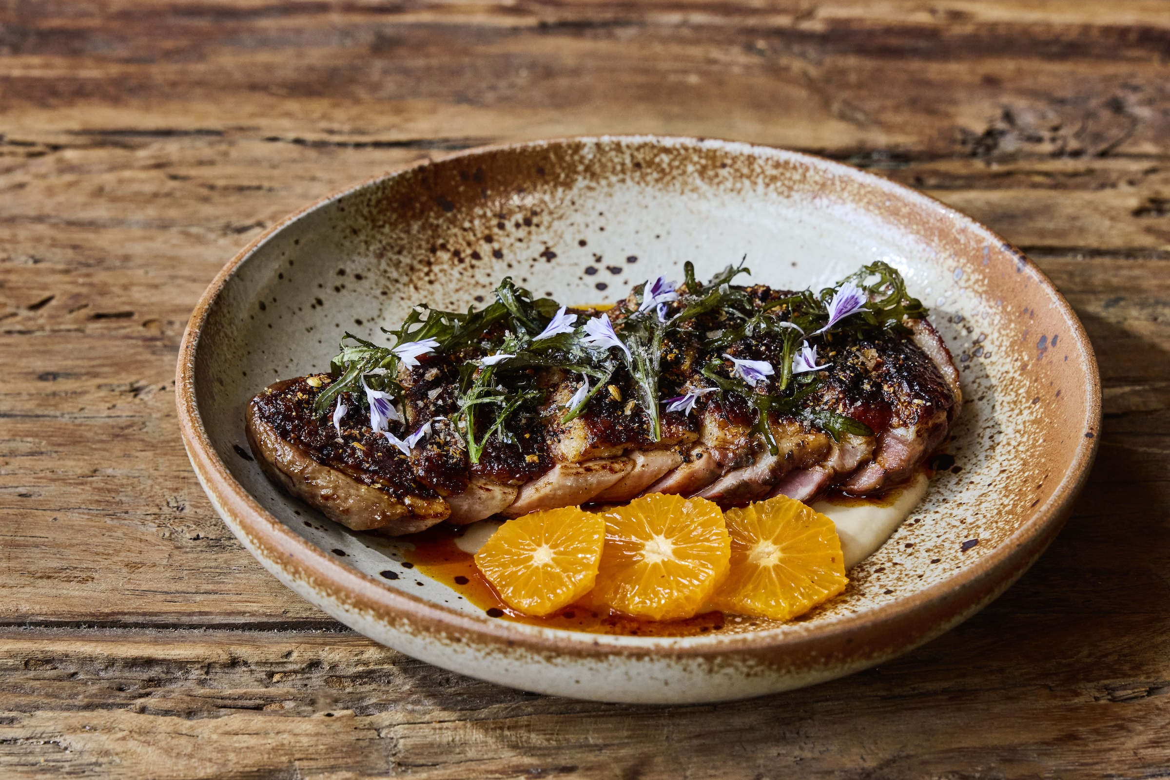 A seared duck breast garnished with greens and edible flowers, served with orange slices on a rustic ceramic plate.